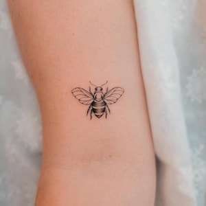May include: A small, black and white bee tattoo. The bee is detailed with striped abdomen and delicate wings. The tattoo is on a light skin tone.