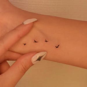 Può includere: Un piccolo tatuaggio temporaneo che presenta quattro sagome nere di uccelli in volo, disposte in una linea orizzontale sulla pelle. Gli uccelli sono raffigurati con linee semplici, con le ali spiegate come se stessero volando.