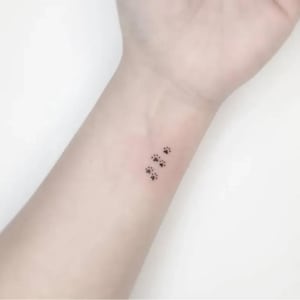 May include: A small, minimalist tattoo on the inner wrist featuring three tiny, black ink paw prints arranged in a diagonal line. Each paw print consists of a central pad and four smaller toe pads, rendered in a simple, clean, and delicate style.