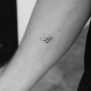 Puede incluir: Un pequeño tatuaje negro de la letra "B" en cursiva en el brazo de una persona. El tatuaje es delicado y parece recién hecho, con líneas finas y un ligero brillo. La piel alrededor del tatuaje es lisa y pálida, contrastando con la tinta oscura.