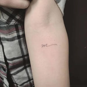 Op de afbeelding: Een kleine, delicate tatoeage van de letter "m" in een cursief schrift. De tatoeage bevindt zich op de binnenarm, met een lichte huidskleur. Het ontwerp is minimalistisch en elegant, met fijne lijnen.