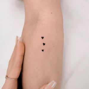 Può includere: Tre piccoli tatuaggi a forma di cuore nero in una linea verticale sul braccio. La pelle è chiara e una mano con un anello è visibile in primo piano.
