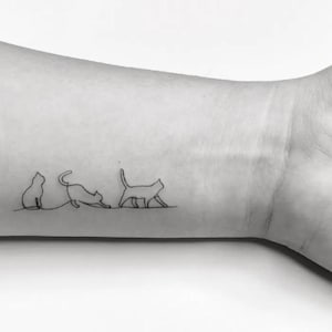 May include: A black ink tattoo on skin featuring a continuous single-line drawing of three cats in different poses. The first cat is sitting, the second is stretching, and the third is walking, all connected by a thin, horizontal line.