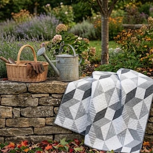 May include: A woven wicker basket with gardening tools and brown leather gloves sits next to a galvanized metal watering can. A folded quilt with a geometric gray and white pattern rests on a stone wall in a garden setting.