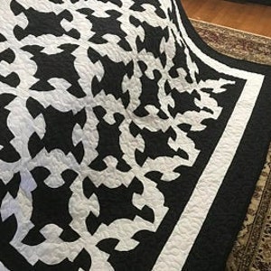 May include: A quilt featuring a high-contrast black and white interlocking pattern. The design consists of repeating white shapes set against a solid black background, finished with a wide black border and intricate overall quilted stitching.
