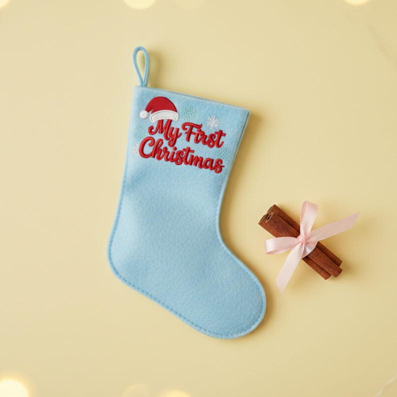 以下が含まれることがあります： 「My First Christmas」の刺繍とサンタ帽が施された水色のクリスマスストッキング。ピンクのリボンで結ばれたシナモンスティックの束がストッキングの隣にあります。ストッキングには吊り下げ用のループが付いています。