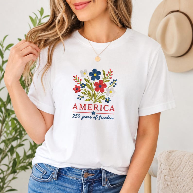 Pode incluir: Camiseta branca com um design floral em vermelho, branco e azul, com a palavra "AMERICA" e o texto "250 years of freedom". A camiseta &eacute; de estilo gola redonda cl&aacute;ssica.