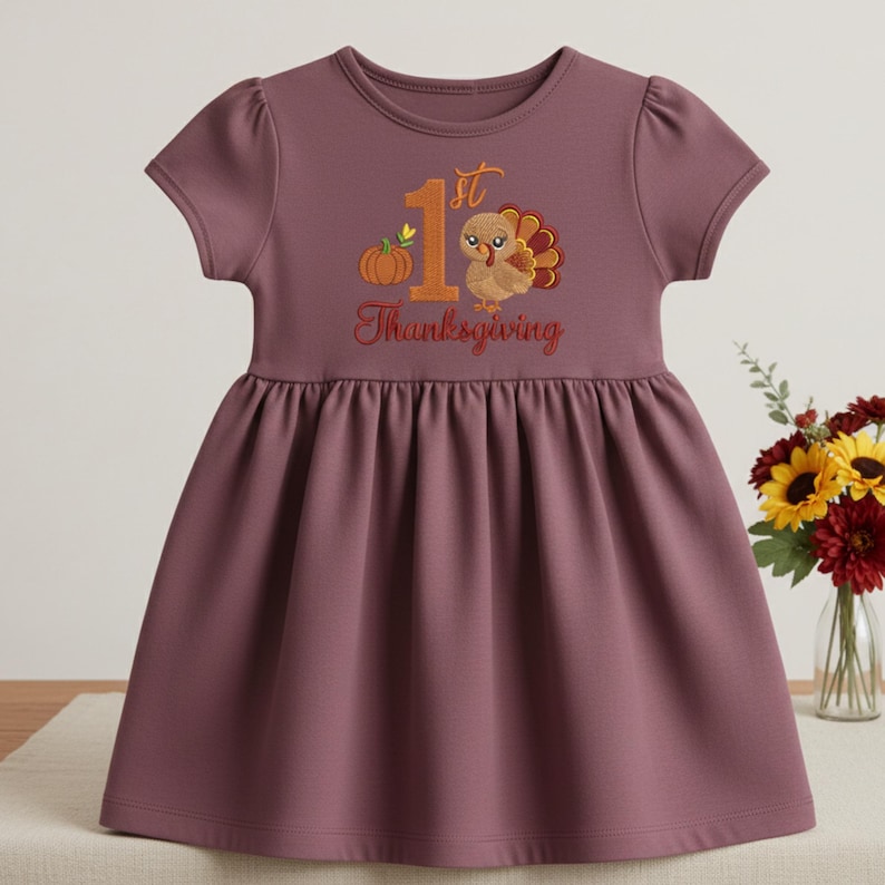 May include: A mauve short-sleeved dress featuring an embroidered graphic of a pumpkin, a turkey, and the text 1st Thanksgiving. The dress has a round neckline, gathered puff sleeves, and a pleated skirt that falls from a high waistline.