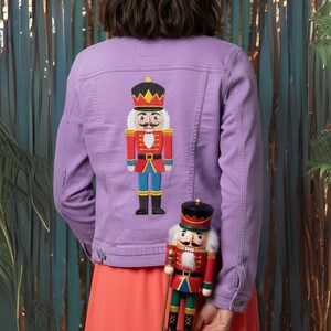 May include: A lavender denim jacket with a colorful embroidered nutcracker design on the back. A matching nutcracker figurine is held in front of the jacket. The jacket has button closures and pockets.