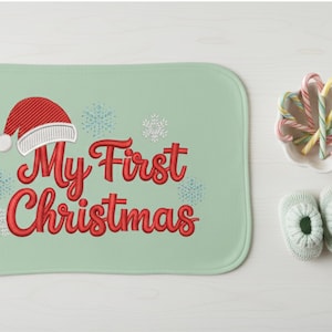 以下が含まれることがあります： 「My First Christmas」の赤い文字が刺繍された、薄緑色のベビーブランケット。サンタ帽と雪の結晶も描かれています。キャンディケインの小皿とベビーブーツも写っています。