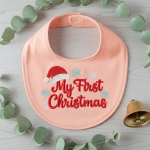 以下が含まれることがあります： 桃色のベビービブ。赤い文字で「My First Christmas」と刺繍されています。赤いサンタ帽子と雪の結晶がテキストの上と周りに刺繍されています。金色のベルとユーカリの小枝が背景にあります。