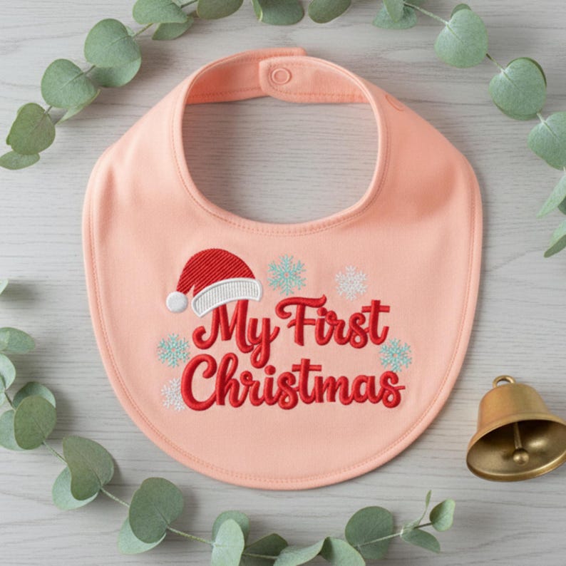 以下が含まれることがあります： 桃色のベビービブ。赤い文字で「My First Christmas」と刺繍されています。赤いサンタ帽子と雪の結晶がテキストの上と周りに刺繍されています。金色のベルとユーカリの小枝が背景にあります。