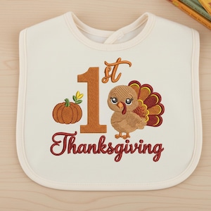 May include: An off-white baby bib featuring an embroidered design with a large orange number 1, the text 1st Thanksgiving in dark red, a small orange pumpkin with green leaves, and a brown turkey with a red and yellow tail.