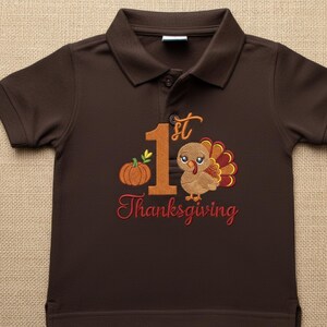 May include: A dark brown short-sleeved polo shirt featuring an embroidered design with a large orange number 1, a small pumpkin, and a turkey. The text 1st Thanksgiving is embroidered in orange and red thread across the front of the shirt.