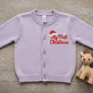 以下が含まれることがあります： ボタン留めのベビー用ライラックカーディガン。セーターには「My First Christmas」の刺繍とサンタ帽のグラフィックが入っています。小さなぬいぐるみトナカイがセーターの隣にあります。