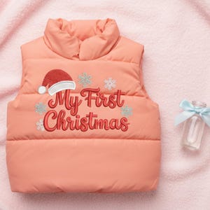 以下が含まれることがあります： 「My First Christmas」の刺繍が入ったコーラル色のパファージャケット。サンタ帽と雪の結晶のデザインが特徴です。右側には青いリボンが付いた小さなガラス瓶があります。