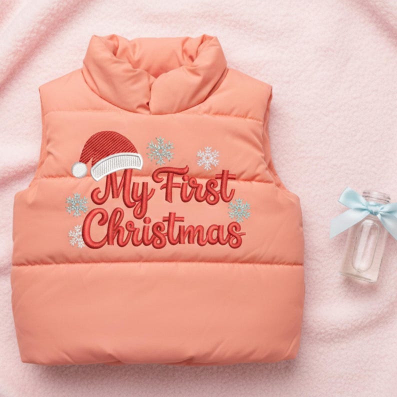 以下が含まれることがあります： 「My First Christmas」の刺繍が入ったコーラル色のパファージャケット。サンタ帽と雪の結晶のデザインが特徴です。右側には青いリボンが付いた小さなガラス瓶があります。