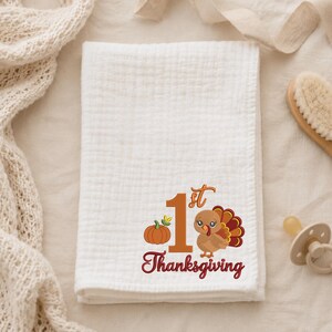 May include: A white muslin swaddle blanket featuring an embroidered design with a large orange number 1, a small orange pumpkin, a cartoon turkey with brown and red feathers, and the red text 1st Thanksgiving printed below the graphic.