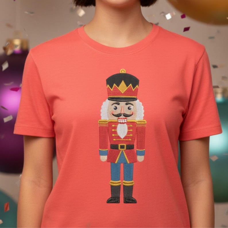 May include: Coral-colored t-shirt featuring an embroidered image of a traditional nutcracker soldier. The nutcracker is depicted in red, black, gold, blue, and white, with a detailed design. The shirt has short sleeves and a crew neck.