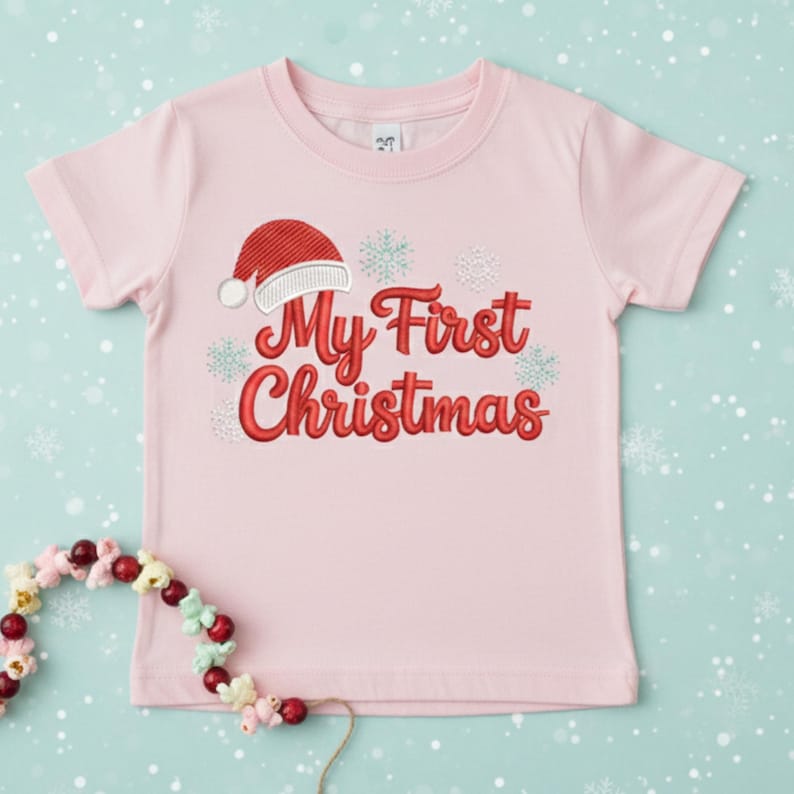 以下が含まれることがあります： 赤い文字で「My First Christmas」と刺繍された、薄いピンク色のTシャツ。赤いサンタ帽子と雪の結晶がテキストの上と周りに刺繍されています。このシャツは半袖です。