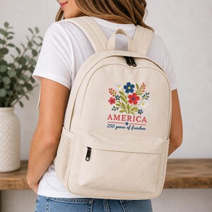 Pode incluir: Mochila bege com um design floral em vermelho, branco e azul, e a palavra "AMERICA" com o texto "250 years of freedom". A mochila tem um bolso frontal e um bolso lateral de malha.