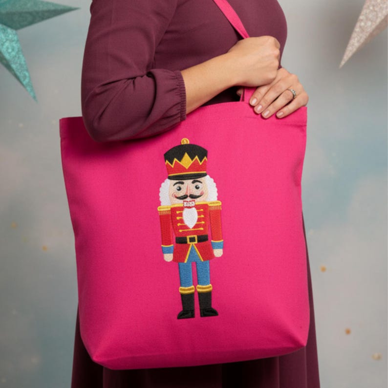 May include: A bright pink tote bag featuring an embroidered image of a traditional nutcracker soldier. The nutcracker is dressed in a red jacket, blue pants, and a black hat. The bag has matching pink handles.