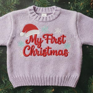 以下が含まれることがあります： 「My First Christmas」の刺繍が入った、薄紫色のニットセーター。赤いサンタ帽と雪の結晶のデザイン。クルーネックのセーターです。