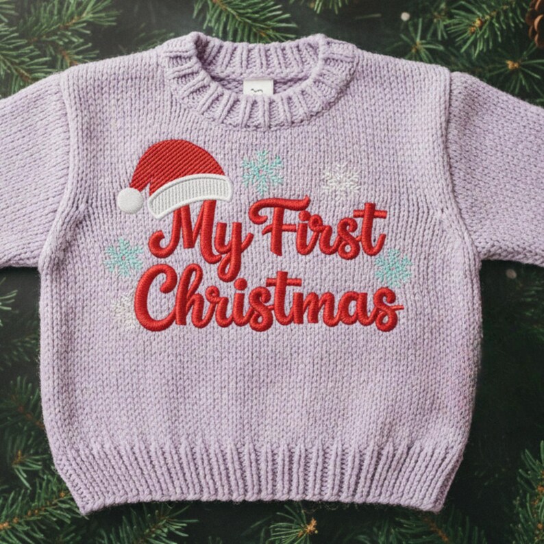 以下が含まれることがあります： 「My First Christmas」の刺繍が入った、薄紫色のニットセーター。赤いサンタ帽と雪の結晶のデザイン。クルーネックのセーターです。