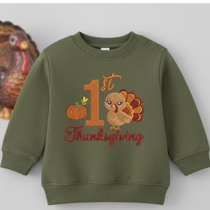May include: An olive green long-sleeve sweatshirt featuring an embroidered design with a large orange number 1, a small pumpkin, and a turkey. The text 1st Thanksgiving is stitched in orange and red thread. The crewneck has two shoulder buttons.