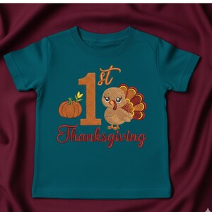 May include: A teal short-sleeved t-shirt featuring an embroidered design with the text 1st Thanksgiving. The graphic includes a large orange number one, a small orange pumpkin with green leaves, and a brown turkey with colorful tail feathers.