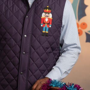May include: A quilted, dark purple vest with a diamond pattern. The vest features a colorful embroidered nutcracker design on the front. The nutcracker is wearing a red jacket, blue pants, and a gold crown. The vest has a zipper and snap closures.