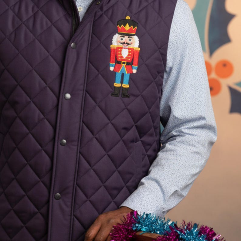 May include: A quilted, dark purple vest with a diamond pattern. The vest features a colorful embroidered nutcracker design on the front. The nutcracker is wearing a red jacket, blue pants, and a gold crown. The vest has a zipper and snap closures.