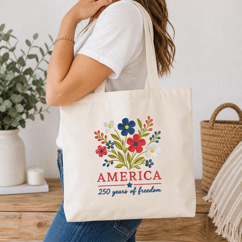 Pode incluir: Bolsa tote creme com um design floral em vermelho, branco, azul e verde. A palavra "AMERICA" est&aacute; impressa em vermelho, com "250 years of freedom" em azul abaixo. A bolsa tem duas al&ccedil;as.