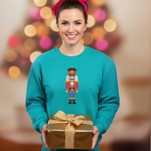 May include: Teal sweatshirt featuring a colorful nutcracker design. The nutcracker is depicted in red, gold, and blue. A gold gift box with a matching bow is held in front of the sweatshirt. The person in the image is wearing a pink headband.