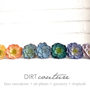 May include: A row of eight faux succulent plants in various shades of yellow, orange, blue, green, purple, and pink. The succulents are arranged in a line on a white surface. The text "DIRT couture" and "faux succulents + air plants + greenery + tropicals" is below the succulents.