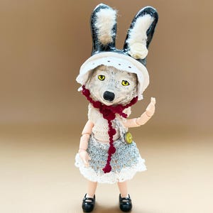 May include: A charming doll with a wolf-like face, wearing a white hat, a cream-coloured vest, and a blue and white skirt. The doll has black shoes and a red beaded necklace. The doll's ears are black with white fur.