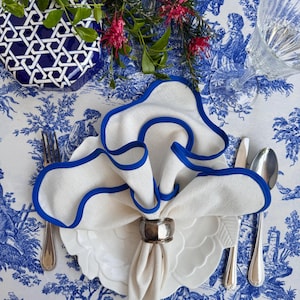 以下が含まれることがあります： 青い縁取りのある白いリネン製のナプキンが、白いプレートの上に置かれ、折り畳まれています。銀製のナプキンリングがナプキンを固定しています。プレートは青と白の模様のテーブルクロスの上に置かれています。