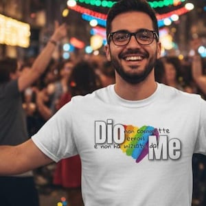 Può includere: T-shirt bianca con la scritta italiana "Dio non commette errori, e non ha iniziato da Me" e un cuore arcobaleno. La persona che indossa la maglietta sorride in mezzo a una folla con luci colorate.