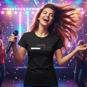 Puede incluir: Una camiseta negra con la palabra "Loading" y una barra de progreso. La persona sostiene un micrófono y está rodeada de gente y confeti en un ambiente brillantemente iluminado.