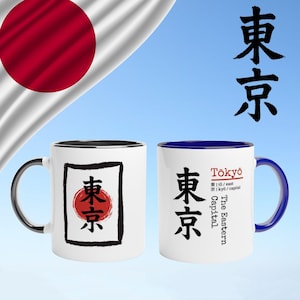 Può includere: Due tazze in ceramica bianca con dettagli neri e blu. Una tazza presenta un cerchio rosso con caratteri giapponesi all'interno di un rettangolo nero. L'altra tazza ha la parola "Tokyo" e caratteri giapponesi. La bandiera giapponese è sullo sfondo.