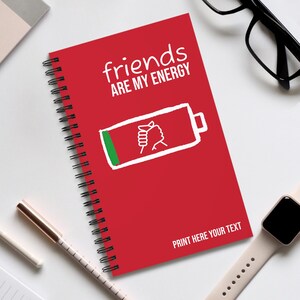 Può includere: Quaderno rosso a spirale con la scritta "friends ARE MY ENERGY" e una grafica a batteria. La grafica a batteria mostra una mano che tiene una spina. La parte inferiore del quaderno recita "PRINT HERE YOUR TEXT".