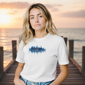 Può includere: T-shirt bianca con grafica skyline blu e la scritta "built to last, strong with pride". Il design è una rappresentazione stilizzata dello skyline di una città. La maglietta è realizzata in materiale morbido.