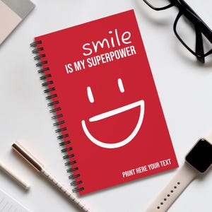 Può includere: Quaderno rosso a spirale con la scritta "smile IS MY SUPERPOWER" e una grande faccina sorridente bianca. La parte inferiore del quaderno recita "PRINT HERE YOUR TEXT". Sono visibili una penna, occhiali e uno smartwatch.
