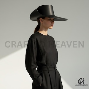Pode incluir: Um chapéu preto com aba larga e um design escultural único. O chapéu é usado com uma camisa preta de manga comprida e calças pretas. O texto "CRAFTED HEAVEN" é visível no fundo.