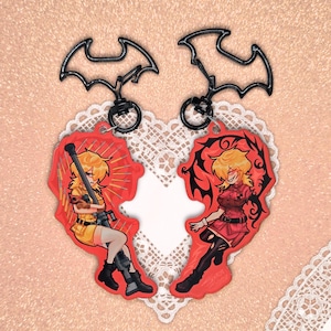 Könnte beinhalten: Zwei Schlüsselanhänger mit Anime-Illustrationen einer Figur in Rot und Gelb. Die Schlüsselanhänger haben die Form eines zerbrochenen Herzens und haben schwarze fledermausförmige Aufsätze. Der Hintergrund ist hellrosa.