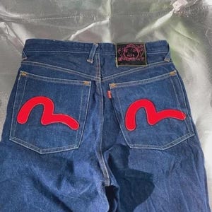 Puede incluir: Vaqueros de mezclilla azul oscuro con dos grandes gráficos rojos en forma de gaviota impresos en los bolsillos traseros. Un parche rectangular de cuero negro con detalles gráficos rosas está cosido en la cintura trasera sobre el bolsillo derecho.