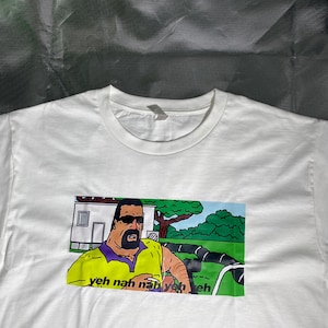 May include: White t-shirt featuring a cartoon image of a man in sunglasses and a yellow shirt. The image includes the text "yeh nah nah yeh yeh eh". The graphic is printed on the front of the shirt.