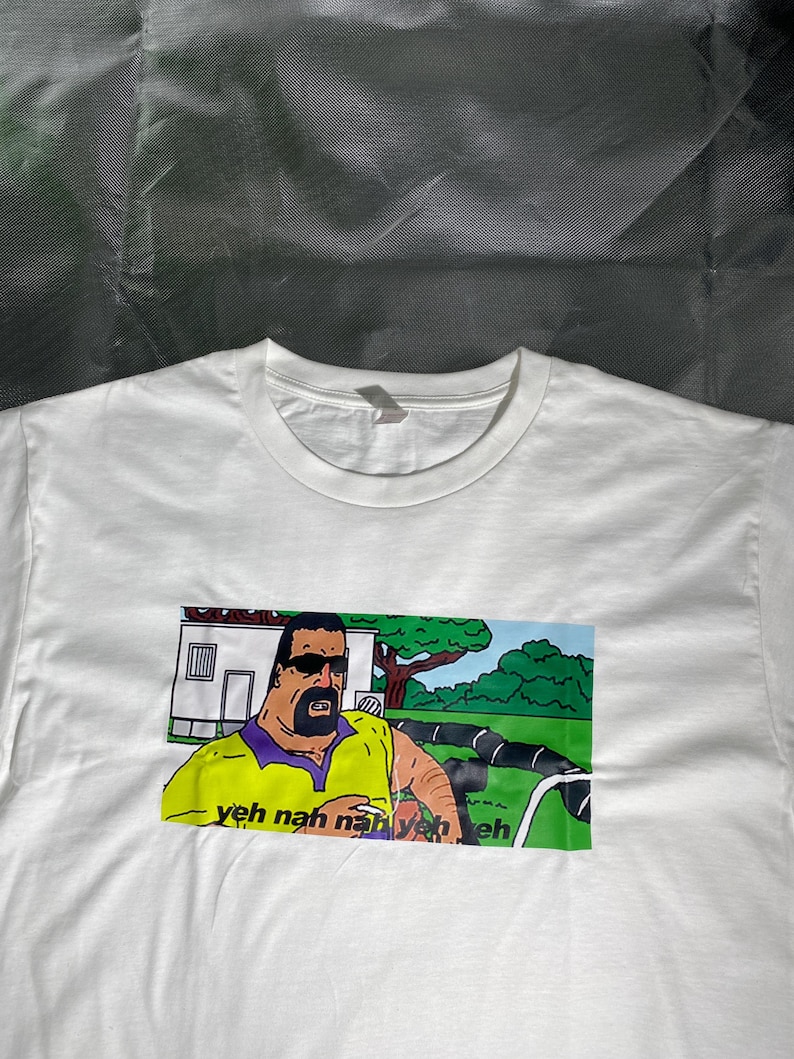 May include: White t-shirt featuring a cartoon image of a man in sunglasses and a yellow shirt. The image includes the text "yeh nah nah yeh yeh eh". The graphic is printed on the front of the shirt.