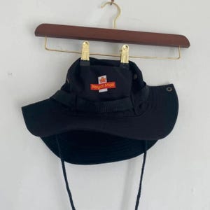 May include: Black bucket hat with a wide brim and a chin strap. The hat features the Royal Mail logo in red and white on the front. The hat is hanging on a wooden hanger.