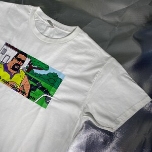 May include: White t-shirt featuring a cartoon image of a man in sunglasses and a yellow shirt. The image includes the text "Nah nah nah yeh eh". The shirt has a crew neck and short sleeves.